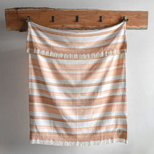 Load image into Gallery viewer, Orange and white striped Escape Throw Blanket with fringes hanging on a wooden rack against a plain wall.